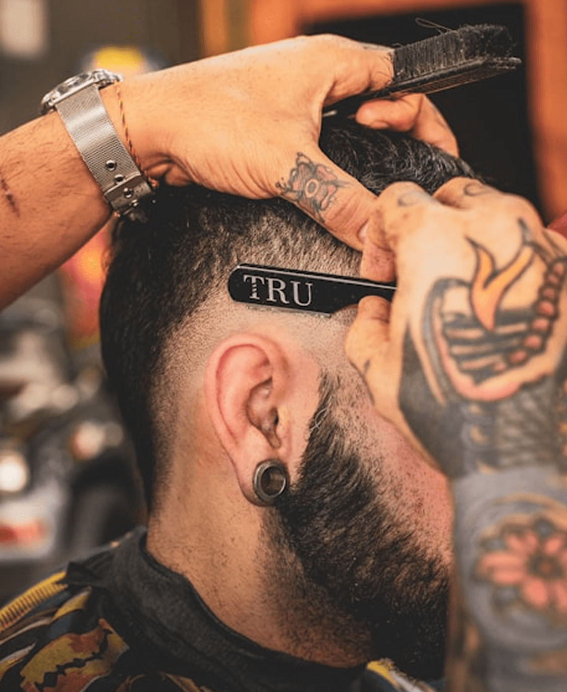 A close-up of a barber using a straight razor to shape a client’s fade haircut. The barber’s hands are tattooed, and the client has a stretched ear piercing.