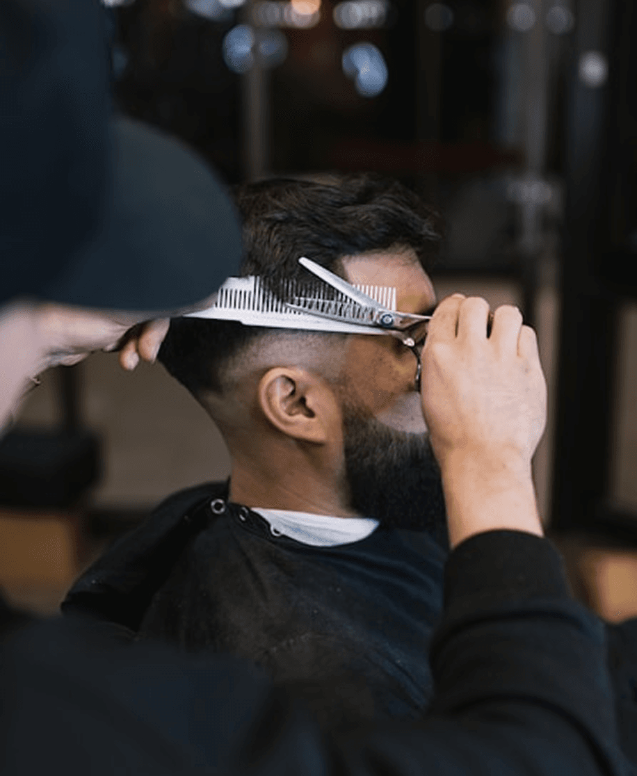 A barber carefully trims a man's hair using scissors and a comb in a barbershop, focusing on the side of the head. The man is draped in a black cape, and the background is softly blurred, showing a barbershop setting with lights and equipment.