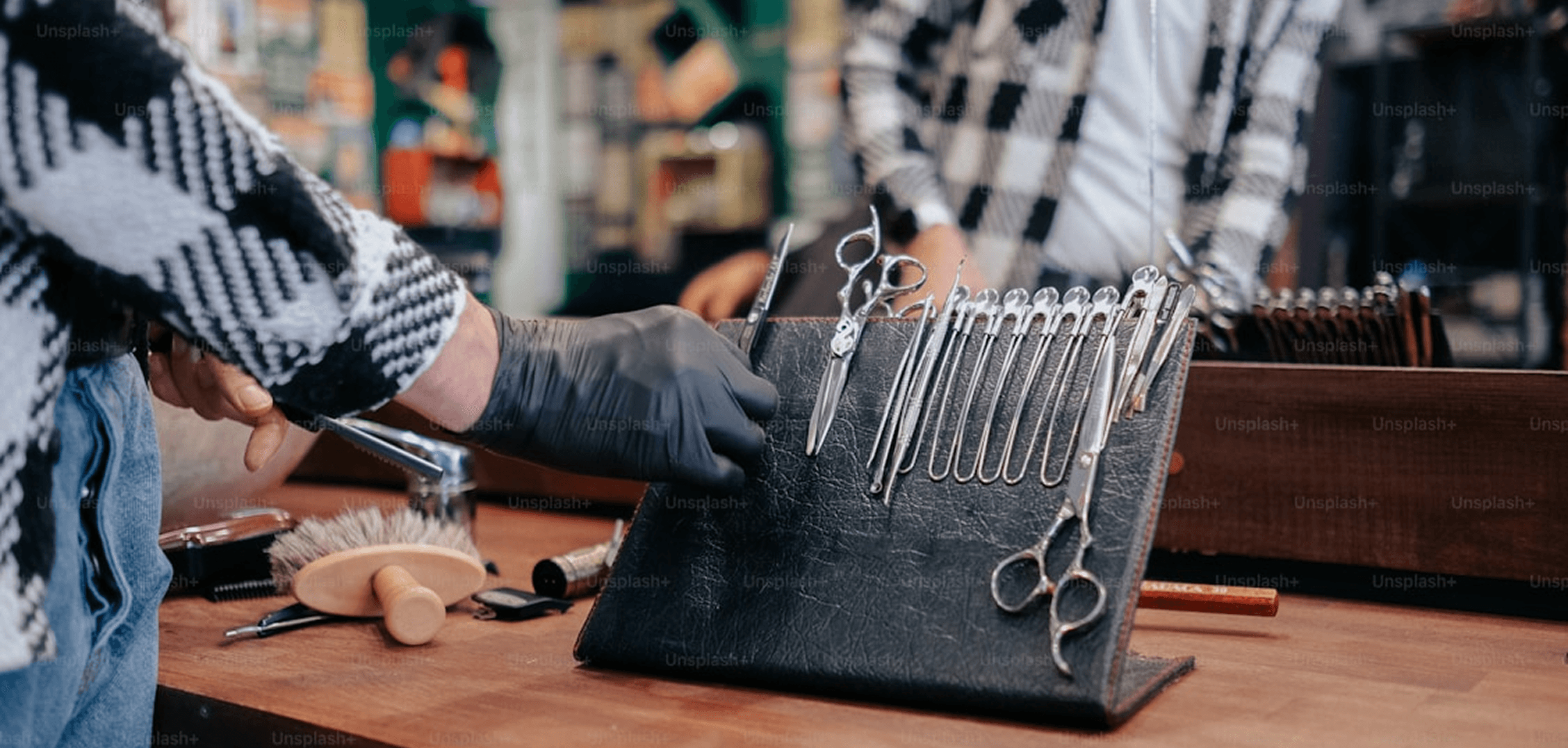 A barber wearing black gloves selects a pair of professional scissors from a leather tool organizer filled with grooming instruments. A mirror and barbershop interior are visible in the background.