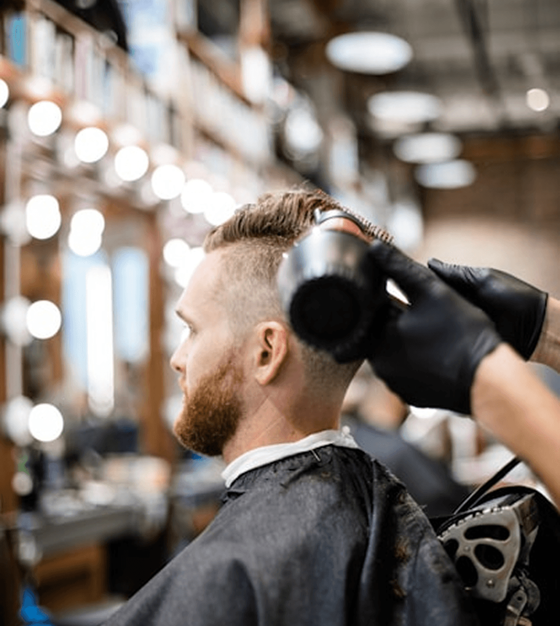 A barber styles a client’s hair using a blow dryer in a modern barbershop. The client has a well-groomed beard and a fade haircut, with a row of bright vanity lights reflecting in the mirror.