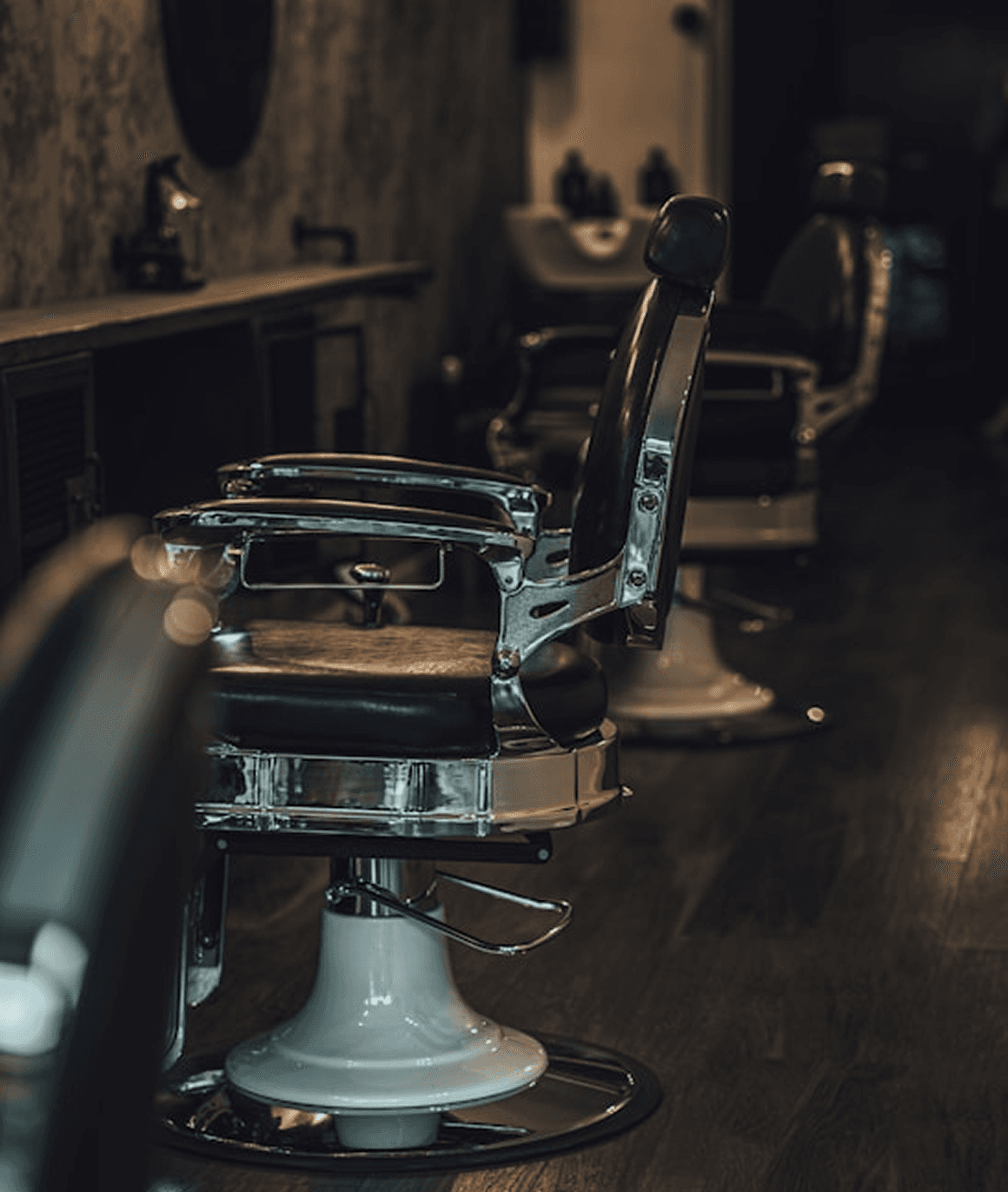 A barber carefully trims a man's hair using scissors and a comb in a barbershop, focusing on the side of the head. The man is draped in a black cape, and the background is softly blurred, showing a barbershop setting.