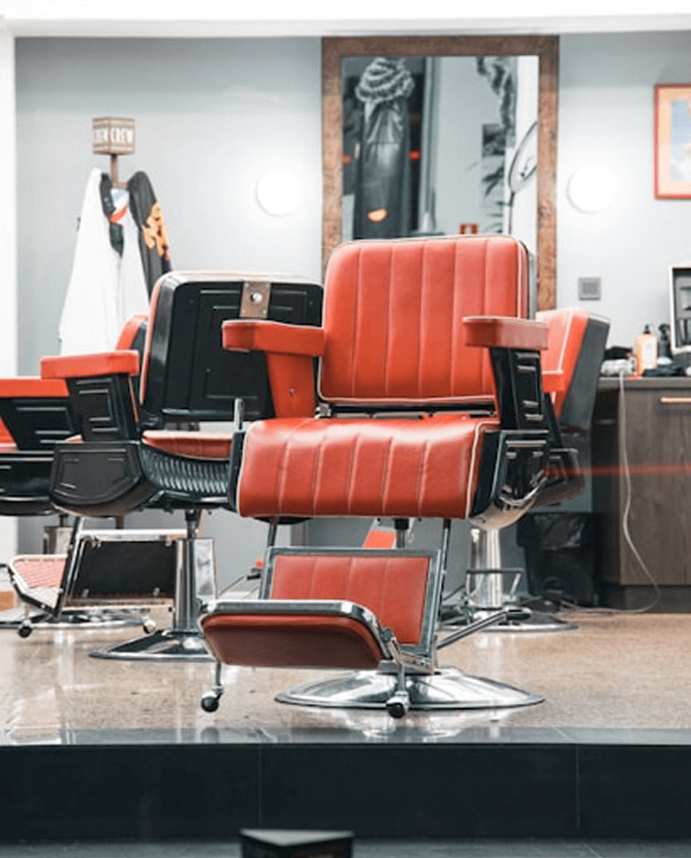 A modern barbershop interior featuring red leather barber chairs with chrome bases, arranged in a row. The space includes mirrors, shelves with grooming products, and a sign reading "True Crew" on the wall, creating a stylish atmosphere.