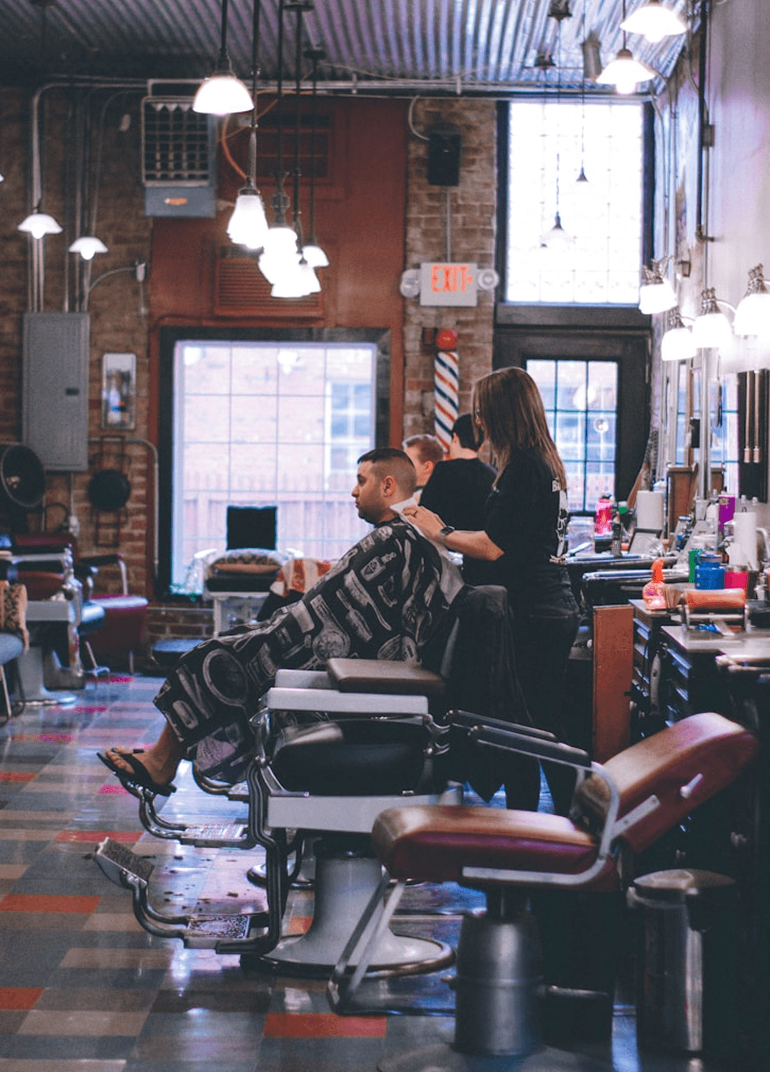 A barber wearing blue gloves trims a client's hair in a barbershop with a rustic brick wall background. The barbershop is decorated with festive garlands and red berries, and features wooden counters with grooming tools.
