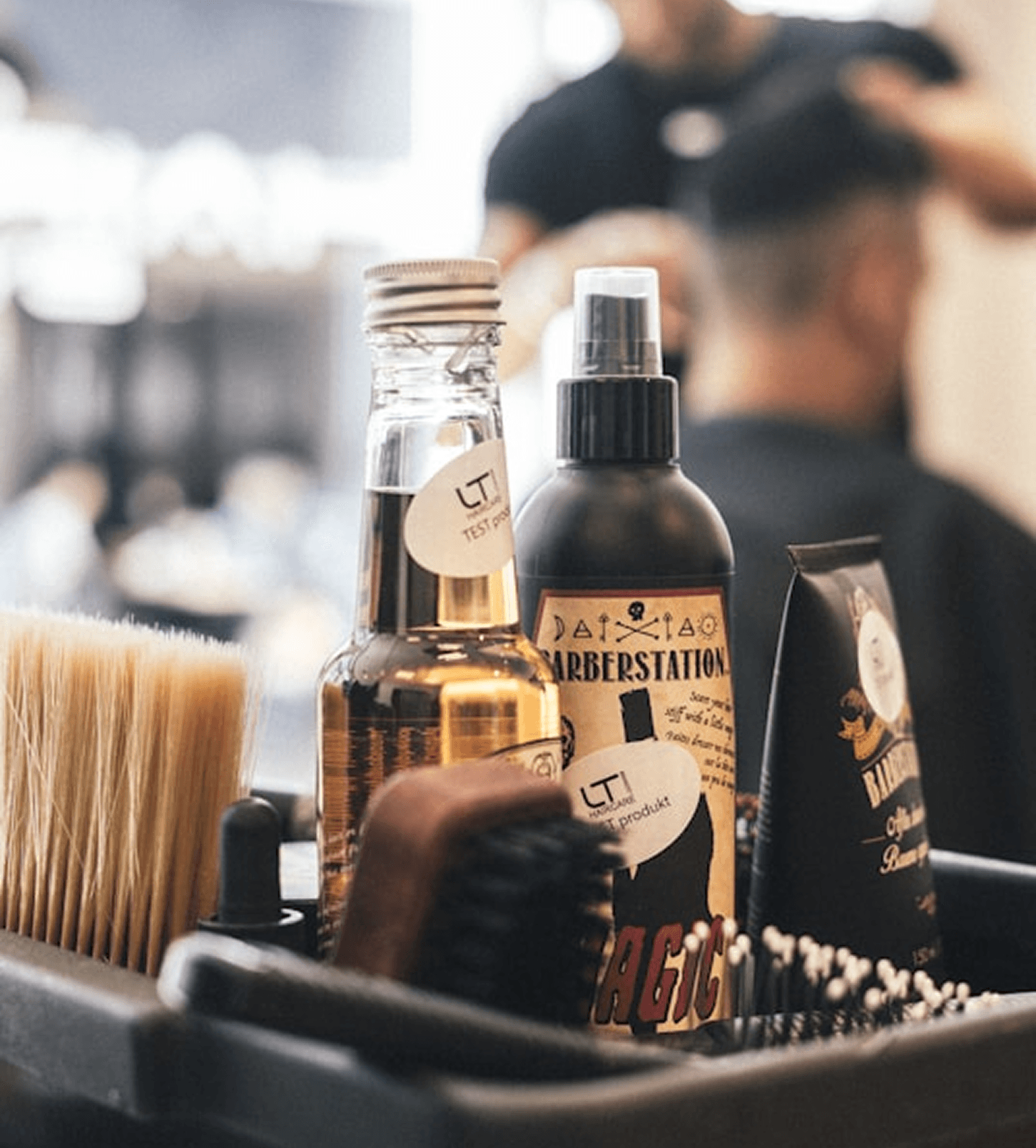 A close-up of barber tools on a counter, including bottles labeled "Barberstation Magic" and "LT Test Product," a brush, and a comb. The background shows a barbershop setting with a client being groomed.