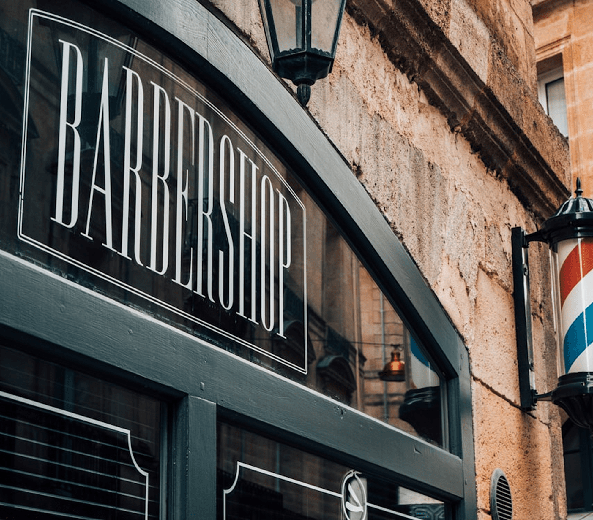 A barbershop interior with a wooden floor and dark walls, featuring multiple barber chairs with black leather upholstery. The space is dimly lit with a vintage aesthetic, including a counter with grooming products and a mirror.