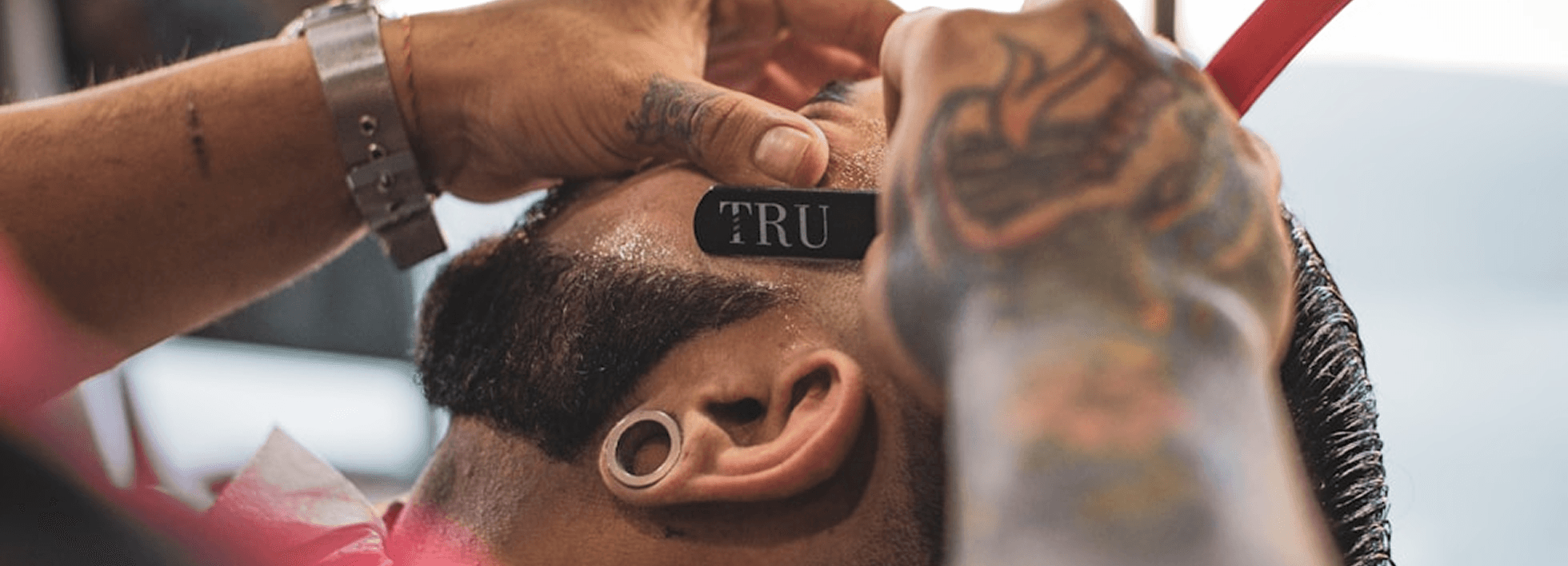 A barber trims a client's hair in a busy barbershop with brick walls and industrial lighting. The client sits in a black barber chair, draped in a patterned cape, while other clients and barbers are visible in the background.